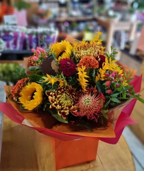 Stunning mixed oranges and reds bouquet with classic orange blooms, celosia, red berries, sunflowers, protea, orange ruscus and complementary flowers. Best of Autumnal Blooms. Bouquet delivered on a aqua wrapping.